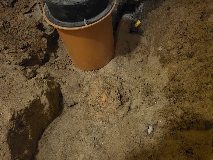 Inspektionslucka i marken med ventil synlig, omgiven av jord och sand. En stor orange rördel finns i bakgrunden.