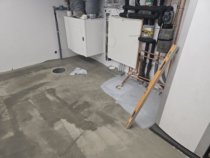 Nyflytspacklat golv i maskinrum med avloppsbrunn och kopparrör på vägg, ojämnt belyst. Avlastningspelare syns, stege lutar mot väggen.