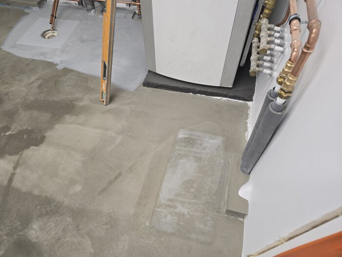 Golvyta under arbete med flytspackel och avstängningslister. Värmepumpsinstallation med kopparrör och synlig inspektionslucka nära golvbrunn.