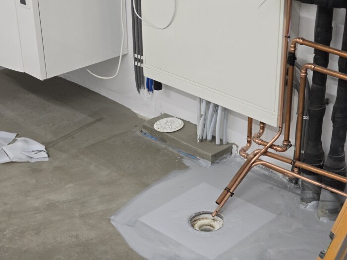 Betonggolv i maskinrum under renovering, med kopparrör och golvbrunn synliga, samt undanlagda verktyg och material i närheten.