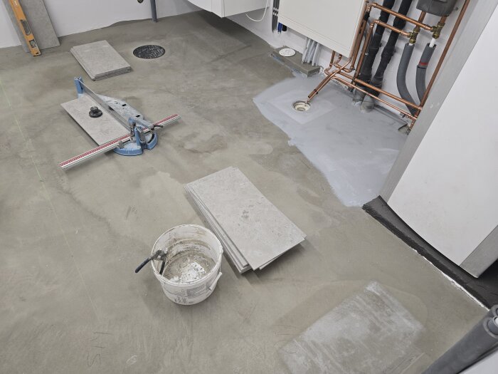 Golv under renovering med flytspackel och plattor, rörinstallation synlig. En kakelskärare och en hink med verktyg finns också på plats.