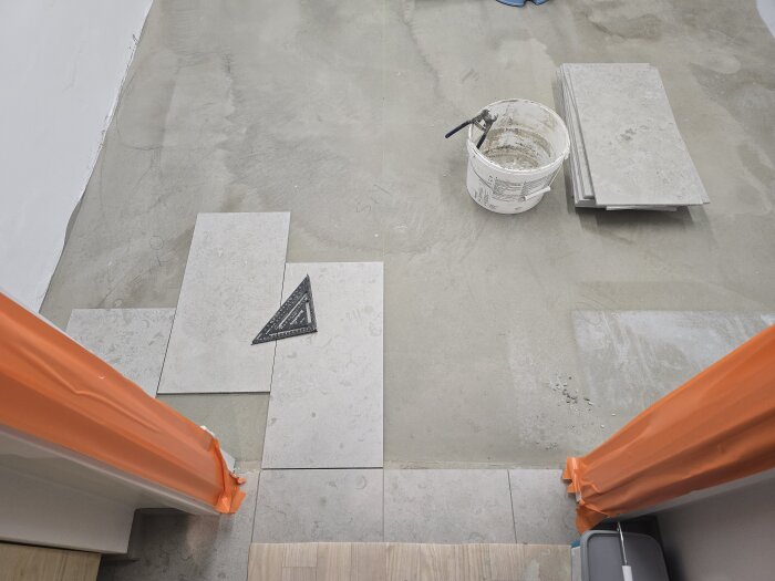 Klinkerplattor läggs ut på ett golv; en murarhink med verktyg syns, och en svart triangel som verktyg ligger bland plattorna.