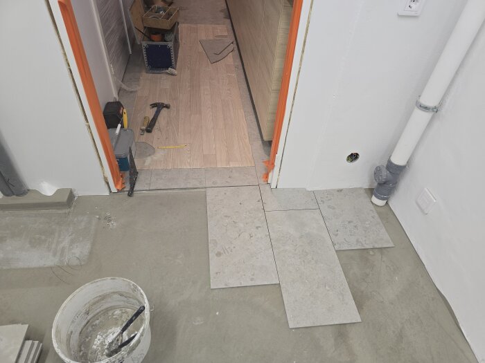 Klinkerplattor ligger utlagda på golvet bredvid en vit vägg och ett rör, med verktyg och betonghink synliga i bakgrunden.