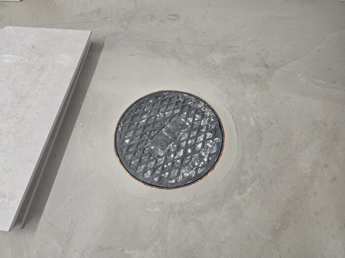 Inspektionslucka i golv med färdigspacklat kakel runtom. En kakelplatta ligger bredvid, redo för montering.