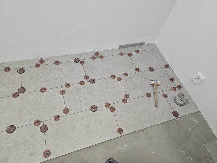 Golvet i ett duschrum under kakelsättning med distanser synliga mellan plattorna. En hammare ligger på golvet nära en inspektionslucka.
