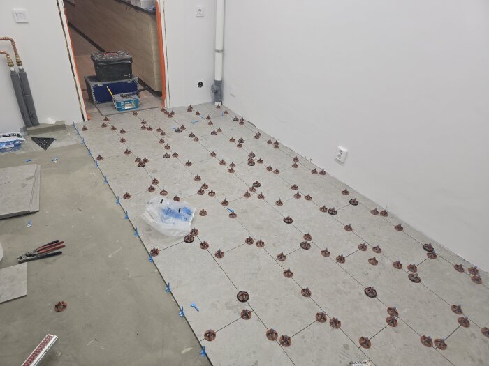 Klinkergolv under installation med nivåjusterande clips i ett rum som ska få ny golvvärme och dörrfoder monterade. Verktyg och material ligger runtom.