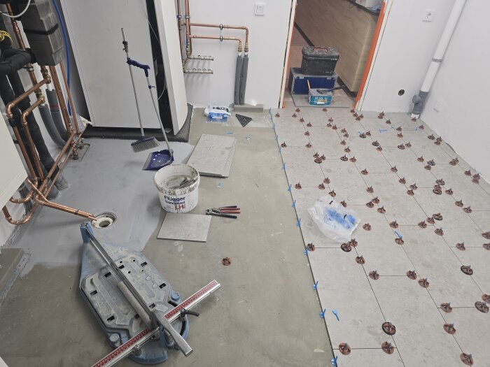 Rum under renovering med golvplattor delvis lagda på golvet, verktyg och material synliga. Rörinstallationer längs väggarna.
