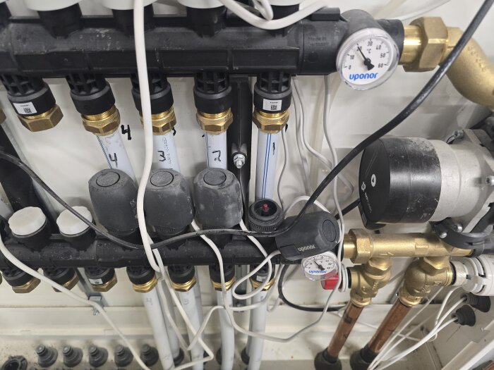 Bild av ett golvvärmesystem med rör, ventiler och en termostat. Ett manometer visar trycket och ledningar syns tydligt.