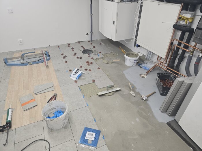 Pågående golvrenovering med klinkerlagda plattor, verktyg och material synliga, samt rörsystem och installationsskåp vid en betongvägg.