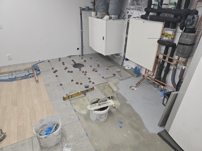 Golvläggning pågår i ett rum; klinkerplattor med distanser, kopparrör, en nivå, och en hink med tools syns. Väggmonterade enheter längst bak.