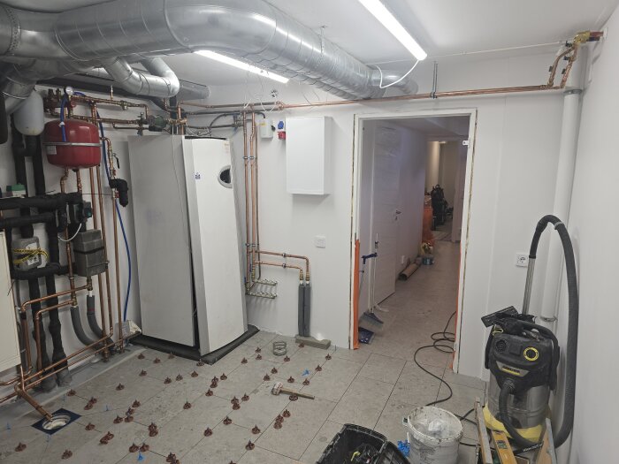 Källarutrymme med golvvärmeinstallation, golvplattor under arbete, rörsystem synliga, dammsugare och verktyg i förgrunden.