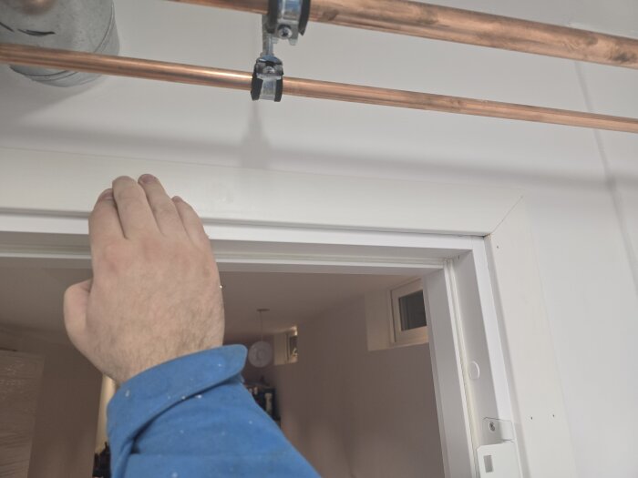 Hand i blå tröja placerad på vitt dörrfoder, med kopparrör ovanför och synlig del av ett rum innanför dörröppningen.