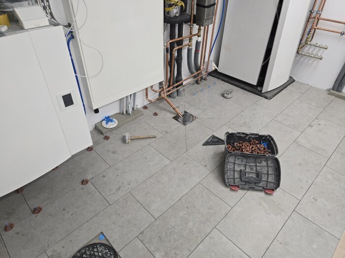 Inspektionslucka på golv med verktyg och arbetsmaterial runtom, kopparrör synliga längs väggen, pågående renovering av våtrum.
