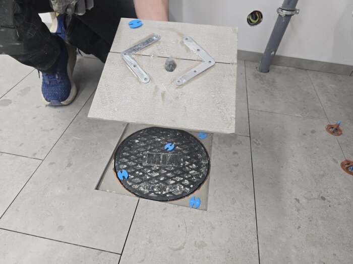 Inspektionslucka på ett kaklat golv lyfts upp, synliggör avrinningsbrunn och rördelar, i ett duschrum under renovering.