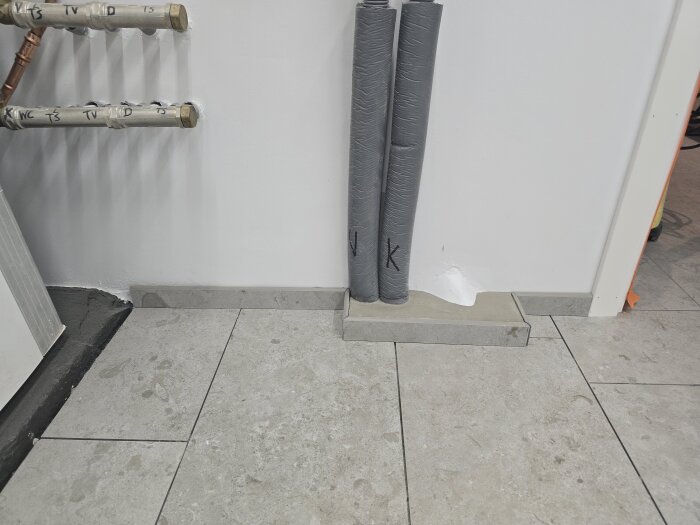 Klinkergolv i duschrum med utskjutande rör och inspektionslucka, rören är isolerade och märkta, väggen är vit och golvet är delvis färdiglagt.