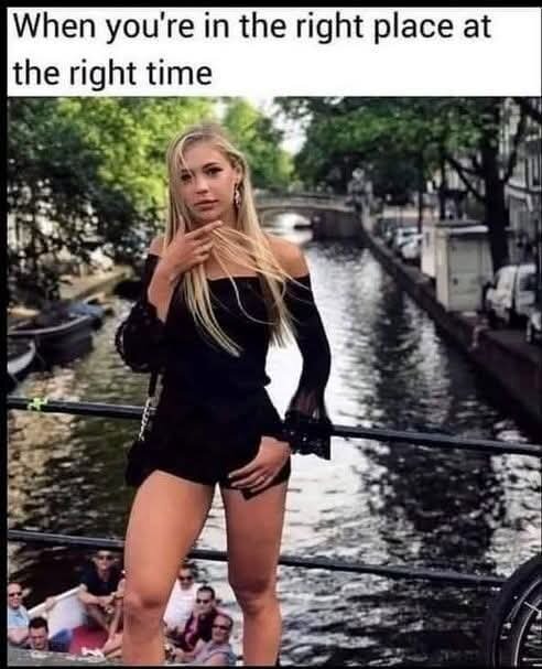 En kvinna i svart klädsel poserar på en bro över en kanal, med gröna träd och båtar i bakgrunden. En grupp människor syns nedanför.