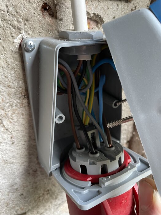 Öppet eluttag med synliga kablar i grå och röd plastkapsling monterat på en betongvägg.