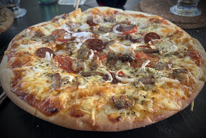 Hemlagad pizza med tomatskivor, porchetta, chorizo, ost, lök, tomatsås, oregano och svartpeppar på ett köksbord.