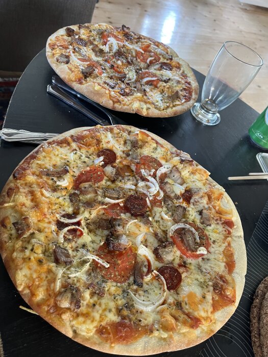 Två hemmagjorda pizzor med tomatskivor, lök, chorizo, porchetta och riven ost på ett svart bord, omgivna av bestick och ett tomt glas.