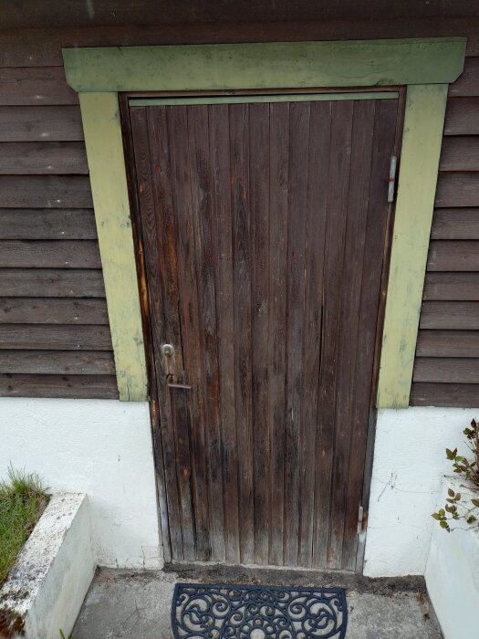 Träfärgad dörr med grönt dörrfoder, sliten yta. Omgiven av brun klädsel och vit fasad. Svart dörrmatta med dekorativt mönster framför.