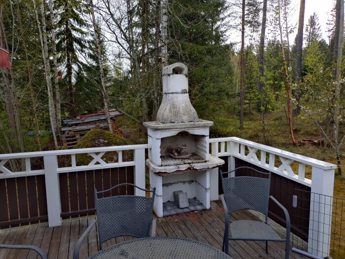 Gammal utegrill i dåligt skick på en altan, omgiven av träd och vedstaplar i bakgrunden.