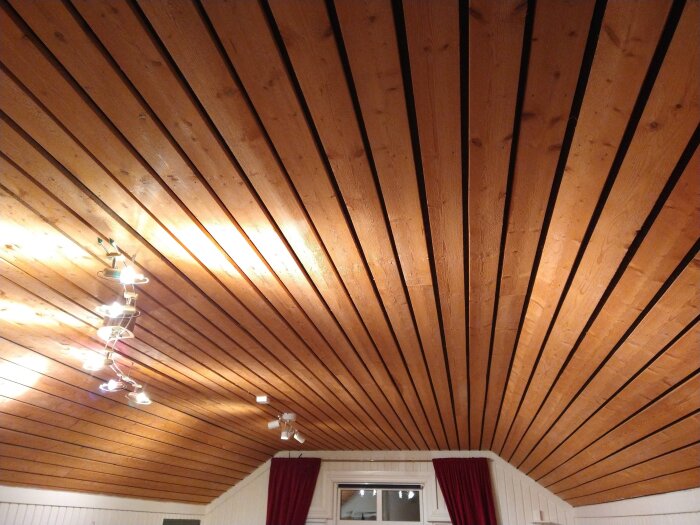 Trätak med bräder som löper från fönstret i mitten utåt, med taklampa till vänster och röda gardiner vid fönstret.