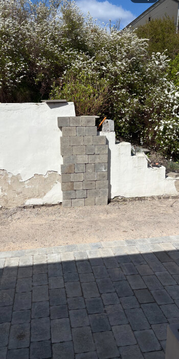 En mur med avskavd puts, delvis täckt med grå betongsten. Bakgrund av buskar med vita blommor.