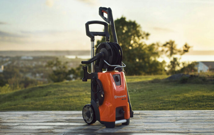 Högtryckstvätt Husqvarna PW360 med orange design och slangvinda, stående utomhus på träaltan med bakgrund av grönskande landskap.