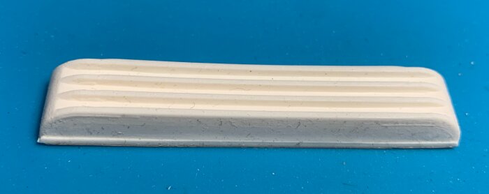 Rektangulär, vit plastbit med rundade hörn och tre upphöjda räfflor på framsidan, cirka 7 cm lång, liggande på blå bakgrund.