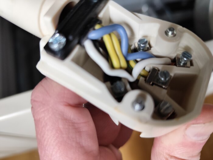 Närbild av stickkontaktens insida med synliga färgade elledningar och skruvar, hålls av en hand.