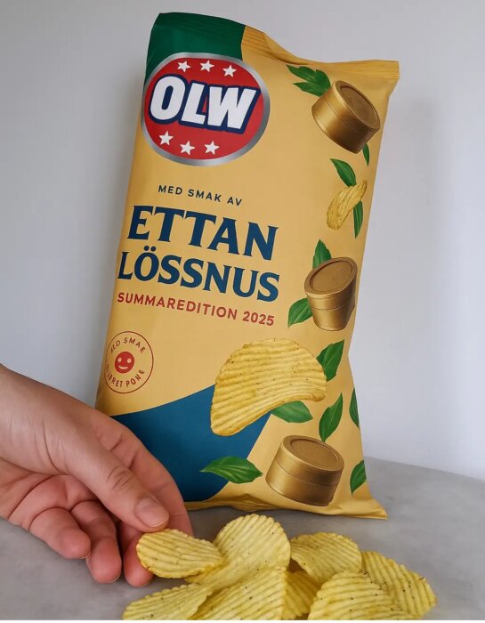 Hand håller en chipspåse med smaken "Ettan Lössnus" från OLW, sommarutgåva 2025, och några chips ligger på bordet.