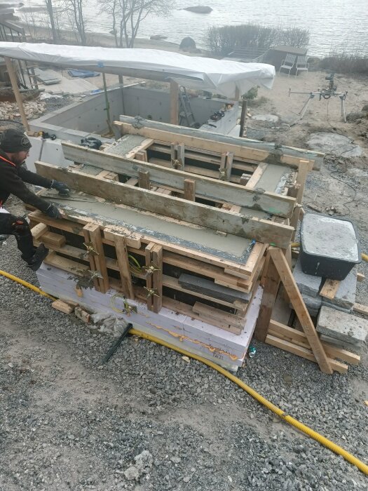En man arbetar vid ett träbeklätt betonggjutningsprojekt nära vatten, med utrustning och material staplade runt omkring.