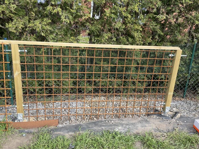 Träram med metallnät upphängd i trädgård, omgiven av gröna buskar.