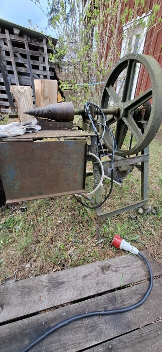 Gammal vedklyvsmaskin med handskar, kablar och en elektrisk anslutning. Står utomhus på träplankor med gräs runtom.