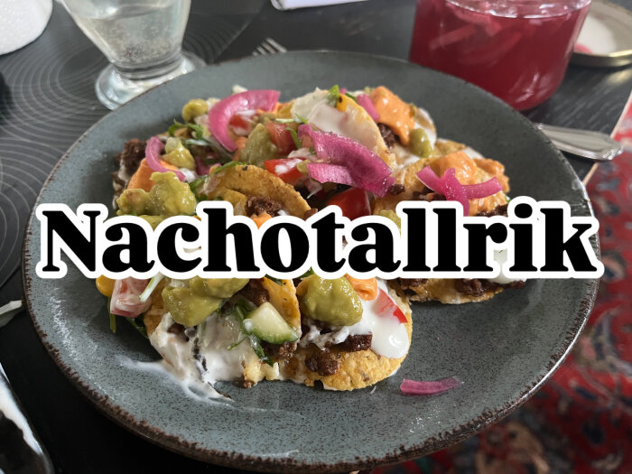 En tallrik med nachos toppade med kött, guacamole, gräddfil, rödlök och olika grönsaker, bredvid en glas med röd dryck.