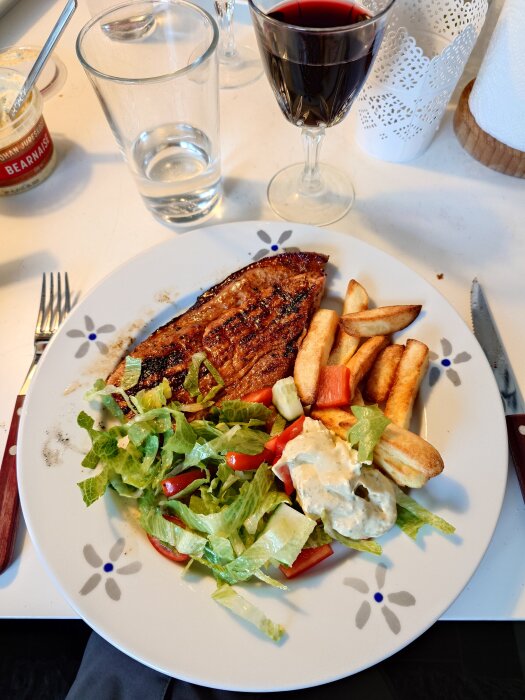 Flintastek på en tallrik med pommes frites, sallad och sås, ett glas rött vin bredvid.