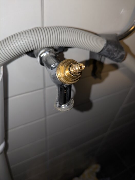 Ventilöverstycke i metall med fastkärvad kägla. En grå slang är fäst vid armaturen som är monterad på en vit kaklad vägg.