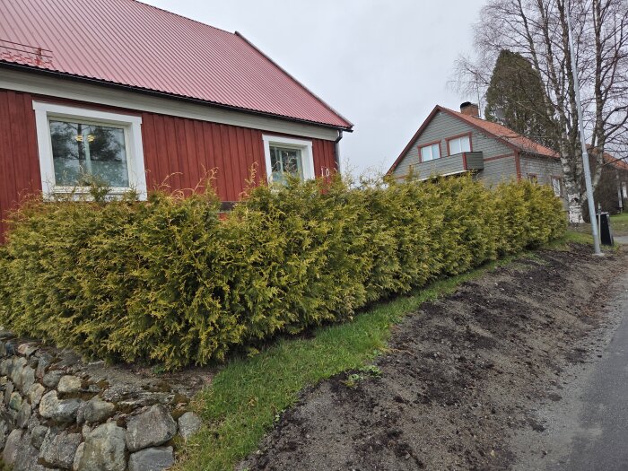 Radhus med röd fasad och häck, gräsyta invid stenmur; frågeställning om gödsling i jordområde vid väg intill.