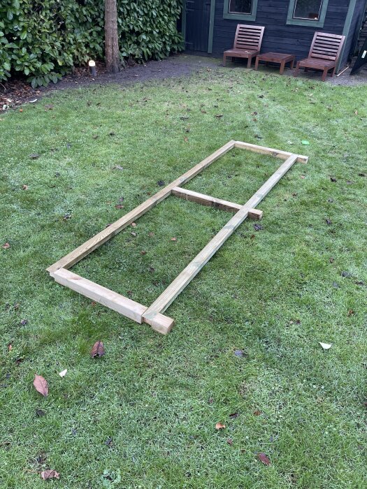 Träram till utomhusbänk på gräsmatta, visar stomme av reglar och stolpar, omgiven av träd och utemöbler.