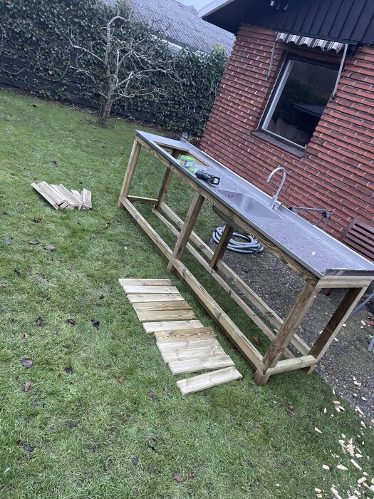 Hemmagjord bänk med rostfri skiva och trästomme på gräsmatta, omgiven av tegelhusvägg och träd. Plankor och verktyg på marken.