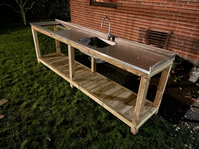 Utomhusbänk med rostfri diskho och avställningsytor, byggd av träreglar och brädor, placerad intill en tegelvägg på gräsmatta.