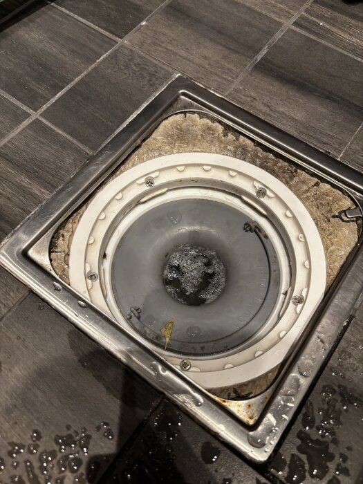 Avlopp med metallklämring i ett renoverat badrum. Grått kakelgolv synligt runtom. Folie och blå manschett är monterade runt brunnen.