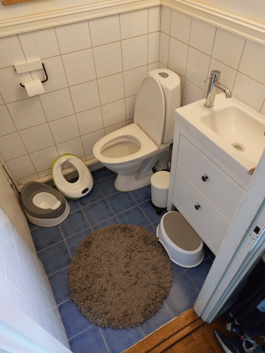 Litet badrum med vit toalett, kommod och handfat. Blått klinkergolv, rund matta och barnpotta. Vit kakelvägg med toalettpappershållare.