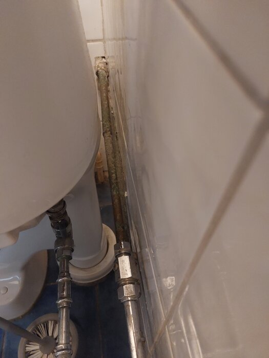 Gammalt rör bakom toalett mot kaklad vägg och blått klinkergolv.