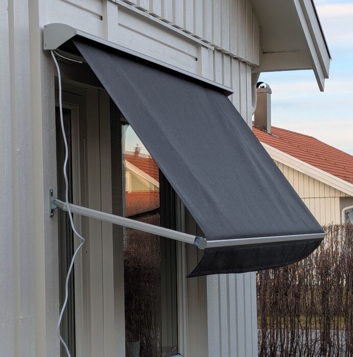 Svart utomhusmarkis monterad på vit träfasad med elkabel, sett från sidan.