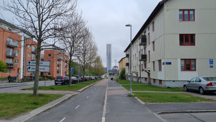 Gata med cykelbana i stadsmiljö, omgiven av bostadshus och parkerade bilar. Ett högt torn syns i bakgrunden.