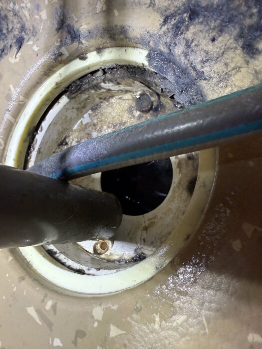 Golvbrunnen i en tvättstuga, med vit plastkonstruktion och synliga rör, omgiven av smuts och vattenrester, sett uppifrån.