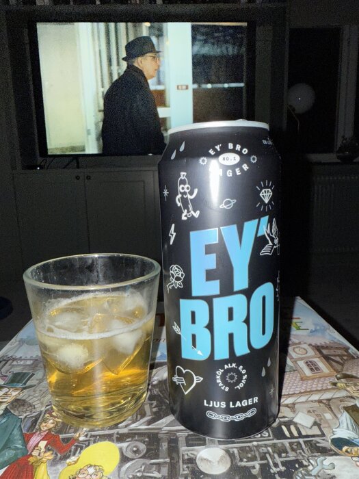 Burk med öl märkt "EY'BRO" bredvid ett glas med isbitar och öl, i bakgrunden syns en TV med en scen där en person går.