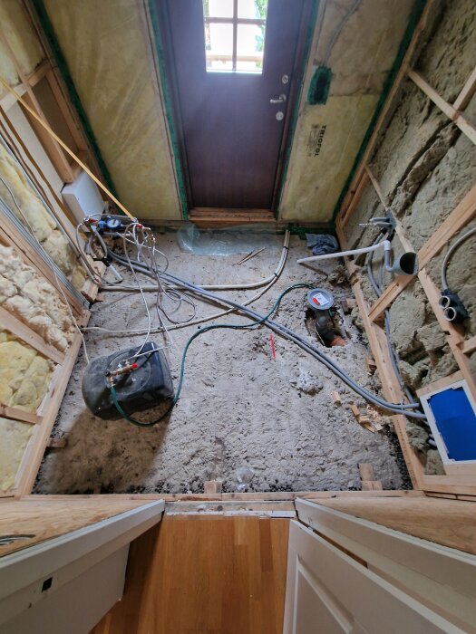 Tvättstuga under uppbyggnad med synliga rördragningar och isolering på väggarna. Betonggolv, trästommar och verktyg synliga.