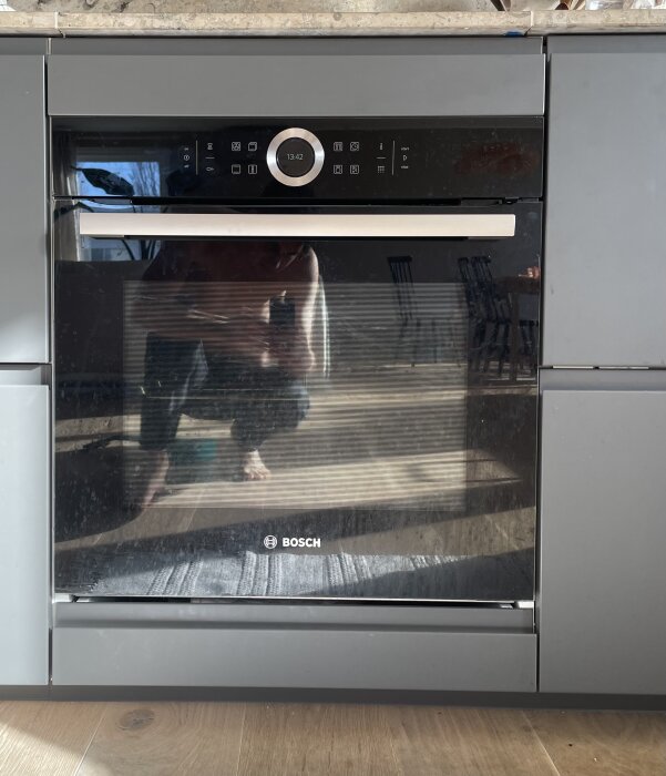 Inbyggd ugn med reflektion av person, tydligt glapp mellan ugn och bänkskiva, synlig metallprofil på skåpsstomme.